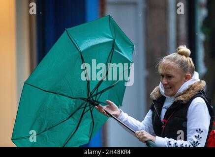 Starker Wind mit Frau, die einen umgekehrten grünen Regenschirm in Preston, Lancashire, Großbritannien, trägt. 1st. November 2021. UK Wetter, kaputter Regenschirm, invertiert, innen heraus, verbogener gebrochener Wind und Regen. Geschäfte, Einkäufer, die an einem nassen und windigen Tag im Stadtzentrum von Preston einkaufen. Quelle: MediaWorlImages/AlamyLiveNews Stockfoto