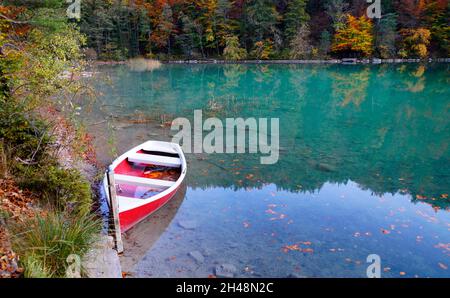 Eine malerische Aussicht auf den Alatsee und ein rotes Boot, das an einem schönen Herbsttag auf dem smaragdgrünen Wasser ruht (Bayern in Deutschland) Stockfoto