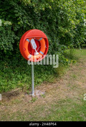 Scampston, North Yorkshire, England, 7. Juli 2021 - leuchtend orange Rettungsring auf einem Metallpfosten mit grünem Laubhintergrund. Stockfoto