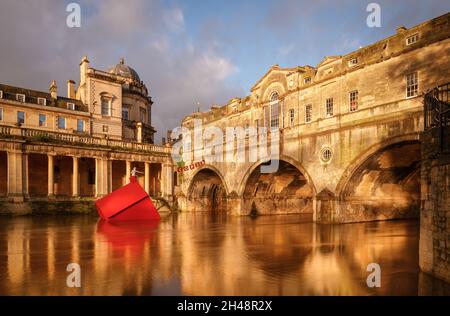 Pulteney Bridge, Bath, 1. November 2021. Bei der Dämmerung in Bath erhellt die Sonne die Pulteney Bridge und die Cop 26-Installation „The Sinking House“, die in Zusammenarbeit mit Stride Treglown, Format Engineers, der Zimmerei Fifield Moss und der Künstlerin Anna Gillespie entworfen wurde. Es ist auch Morgendämmerung bei der Klimakonferenz der Vereinten Nationen, wo die Staats- und Regierungschefs der Welt sich mit Fragen einer erhöhten Bedrohung in der sich verschärfenden globalen Klimakrise befassen werden. „The Sinking House“ ist eine Botschaft der Warnung und der Hoffnung. Kredit: Casper Farrell/Alamy Nachrichten Stockfoto