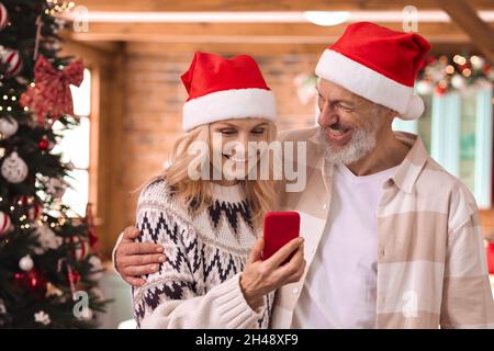 Glückliches 50-jähriges Paar in weihnachtsmützen, das zu Weihnachten zu Hause mit dem Handy telefoniert. Stockfoto