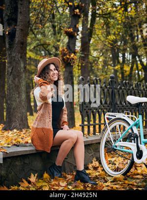 Porträt von niedlichen weiblichen lockigen Biker, die im Herbst Park mit Retro-City-Zyklus ruhen. Wandern im Freien mit dem Fahrrad. Schöne Erholung beim Radfahren bei frischem Herbstwetter. Stockfoto