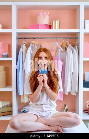Kaukasisch schön aussehende schöne fröhliche Dame mit Kreditkarte in den Händen, macht Online-Shopping, Vorbereitung für die Veranstaltung in hellweißem Innenraum, Station Stockfoto