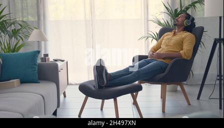 Junger Mann, der sich im Wohnzimmer zu Hause entspannt und Musik hört, sitzt auf einem Sessel und trägt kabellose Kopfhörer Stockfoto