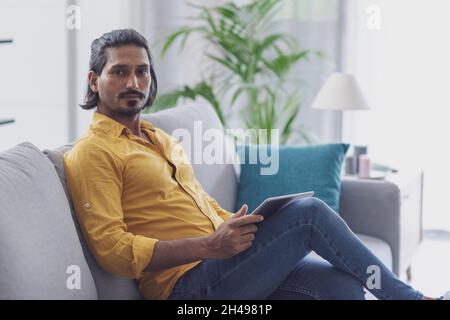 Gutaussehender Mann, der sich zu Hause auf der Couch entspannt und ein digitales Tablet benutzt, schaut auf die Kamera Stockfoto