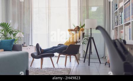 Der indische Mann, der sich zu Hause im Wohnzimmer entspannt und Musik hört, sitzt auf einem Sessel und trägt kabellose Kopfhörer Stockfoto