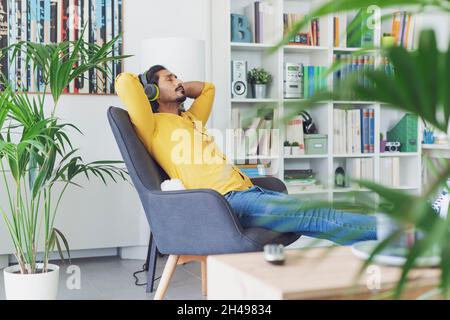 Junger Mann, der sich im Wohnzimmer zu Hause entspannt und Musik hört, sitzt auf einem Sessel und trägt kabellose Kopfhörer Stockfoto