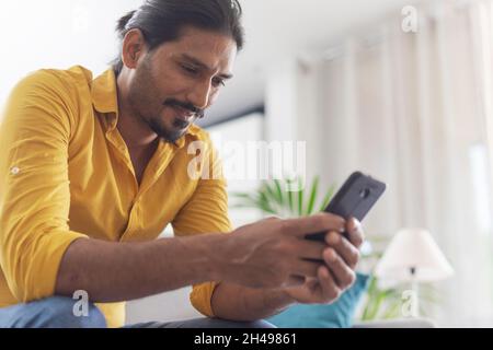 Der junge Mann sitzt zu Hause auf der Couch und chattet mit seinem Smartphone, Kommunikations- und Technologiekonzept Stockfoto