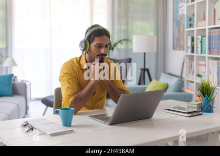 Mann, der zu Hause am Schreibtisch sitzt und sich mit seinem Laptop verbindet, trägt kabellose Kopfhörer und schaut sich online Videos an Stockfoto