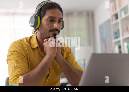 Mann, der zu Hause am Schreibtisch sitzt und sich mit seinem Laptop verbindet, trägt kabellose Kopfhörer und schaut sich online Videos an Stockfoto