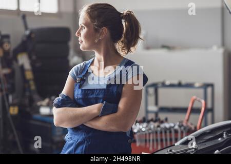 Porträt einer selbstbewussten Mechanikerin, die in der Autowerkstatt posiert, schaut weg und lächelt Stockfoto