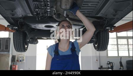 Die Mechanikerin, die unter einem Auto in der Autowerkstatt arbeitet, prüft auf Undichtigkeiten Stockfoto