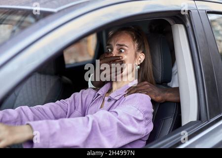 Afrikanischer Mann griff wehrlose kaukasische Frau auf dem Parkplatz an. Vergewaltiger versucht, das Opfer zu entführen. Seitenansicht auf verängstigtes junges Weibchen, das sitzt i Stockfoto