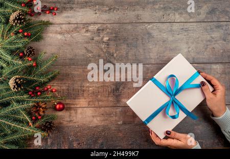 Frau Hände halten weihnachtsgeschenk oder Geschenkbox dekoriert Tannenbaum. Kuscheliger Winteranschlag. Weibliche Hände im Pullover halten Weihnachtsgeschenk-Box auf Holz Stockfoto