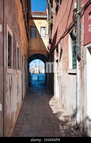 Schmale Straße in Giudecca, Insel in der venezianischen Lagune. Historische Häuser bilden einen Bogengang, der in Richtung Kanal mit blauem Wasser und historischem führt Stockfoto