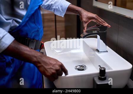 Nahaufnahme der afrikanischen schwarzen Klempnerhand, die versucht, einen Wasserhahn im Badezimmer zu öffnen, repariert ein Handwerker oder Klempner in blauer Uniform die Installation des Wasserhahns Stockfoto