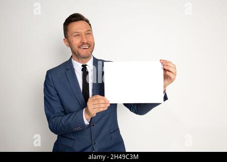 Junger hübscher, lachender Brunet-Mann mit weißem leeren Brett. CEO, Manager, Geschäftsmann, der Plakat für Werbung zeigt. Studio auf weißem Hintergrund gedreht Stockfoto