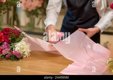 Floristin Frau Schneiden Umschlag für Blumenstrauß Dekoration mit Schere, im Geschäft. Cropped weiblich in Schürze konzentriert auf die Arbeit, sorgfältig sauber machen b Stockfoto