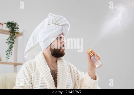 Bärtiger Rüde mit Badetuch auf dem Kopf, der eine Sprühwasserbehandlung auf das Gesicht anwendet. Spa-, Körper- und Hautpflege für den Mann Konzept. Stockfoto
