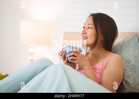 Glückliche asiatische Frau, die im Bett lag und morgens Kaffee trank Stockfoto
