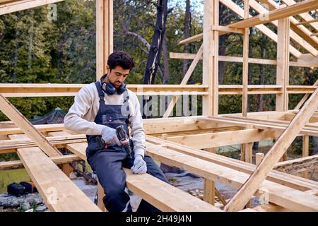 Junge Hart Arbeitende Kaukasische Auftragnehmer Arbeitet Mit Drill Driver. Zimmermann arbeitet an Holzhaus Skelett Rahmen Dachabschnitt. Bauindustrie Stockfoto