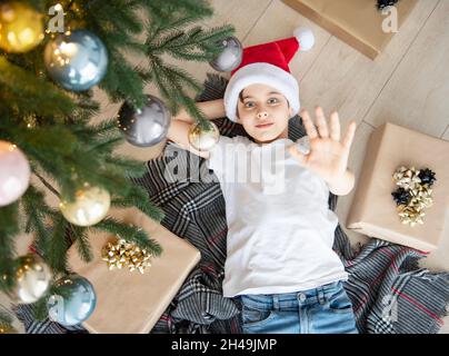 Der Junge liegt unter dem Baum und träumt. Weihnachtsbaum im großen Wohnzimmer. Stockfoto