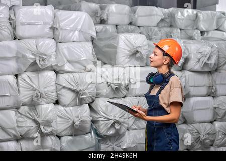 Frau in Hardhat Zählen verpackt chemischen Kunststoff-Behälter in der Fabrik, Notizen machen, hält Klemmbrett Ordner in den Händen, trägt Uniform, vorbereitet Schädling Stockfoto