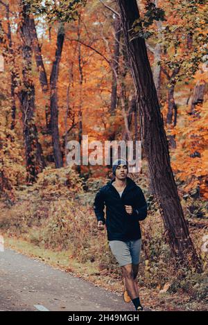 Im Herbst joggt ein gesunder junger, braunhäutiger Mischling auf dem Trail im Wald. Mit Beanie und schwarzer Jacke, die durch den Wald läuft Stockfoto