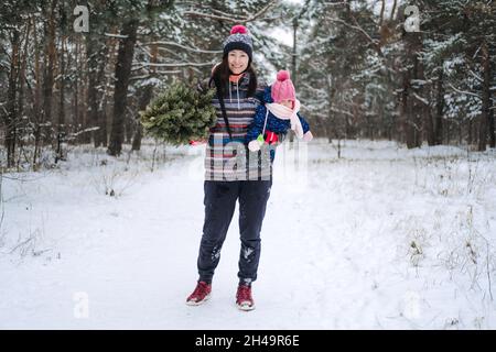 Junge Frau, alleinerziehende Mutter und Kleinkind Baby Mädchen trägt Weihnachtsbaum im Winterpark. Vorbereitung auf Weihnachten, Kommissionierung, Auswahl Weihnachtsbaum. Stockfoto