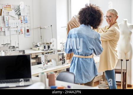 Glückliche Dame mittleren Alters Schneiderin unterrichtet junge afroamerikanische Stagistin in Workshop Stockfoto