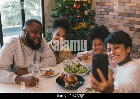 Fröhliche afroamerikanische Familie genießt weihnachten und das Mittagessen im neuen Jahr, während die Frau Selfie auf dem Smartphone mit Familienmitgliedern zu Hause gegen geschmückten Weihnachtsbaum nimmt Stockfoto