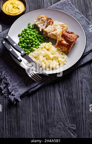 Gebratener Schweinelende und Sauerkraut auf einem Teller mit grünen Erbsen und Senf auf dunklem Holzboden, Draufsicht, Nahaufnahme Stockfoto