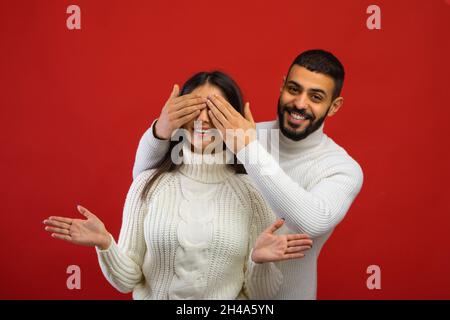 Wer ist es? Positiver arabischer Mann, der die Augen der Frau von hinten bedeckt, hinter ihr steht und die Kamera anlächelt, glückliche Ehegatten, die sich über den roten Studiohinterzimmer lustig machen Stockfoto