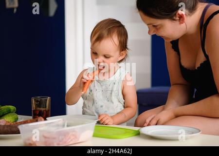 Porträt eines kleinen Mädchens, das eine Wurst isst, ihre Mutter sitzt daneben und beobachtet sie Stockfoto