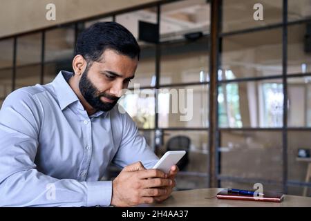Lächelnder indischer ceo Geschäftsmann hält Telefon an einem modernen Schreibtisch. Stockfoto