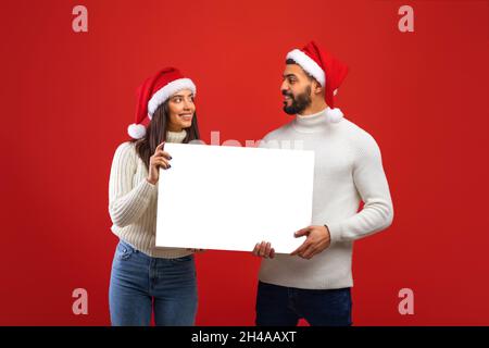 Glückliches arabisches Paar in Santa-Hüten, das weiße Werbetafel hält, leeren Platz für Ihren Text oder Ihr Design zeigt, auf rotem Hintergrund und Smili steht Stockfoto