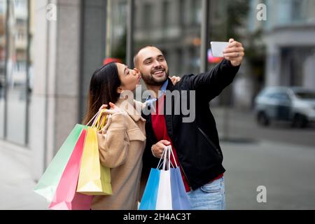 Liebevolles interrassisches Paar mit bunten Taschen, das Selfie auf dem Handy vor dem großen Einkaufszentrum nimmt. Junge, vielfältige Familie fotografiert sich während g Stockfoto