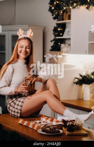 Ein Mädchen sitzt am Weihnachtstag auf dem Küchentisch und hält ein Glas Champagner in den Händen.Eine Frau am Silvesterabend in der Küche mit Champagner. Stockfoto