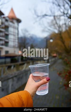 Blick aus der ersten Person mit einem Glas reinem Wasser aus einer Bergquelle vor dem Hintergrund der Stadt Borjomi. Stockfoto