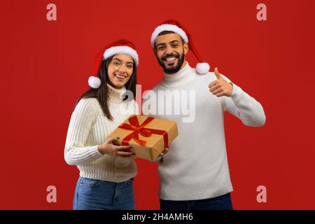 Glückliches, junges arabisches Paar in Weihnachtsmützen mit Geschenkbox und Mann, der mit Daumen nach oben zeigt, posiert und lächelt vor der Kamera auf rotem Studiohintergrund Stockfoto