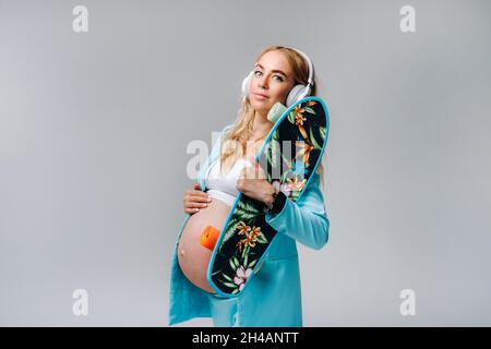 Ein Schwangere im türkisfarbenen Anzug mit Skateboard in den Händen und Kopfhörern steht auf einem grauen Hintergrund. Stockfoto