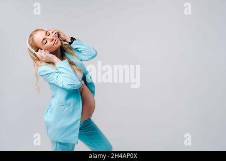 Eine Schwangere im türkisfarbenen Anzug mit Kopfhörern steht und hört Musik auf grauem Hintergrund. Stockfoto