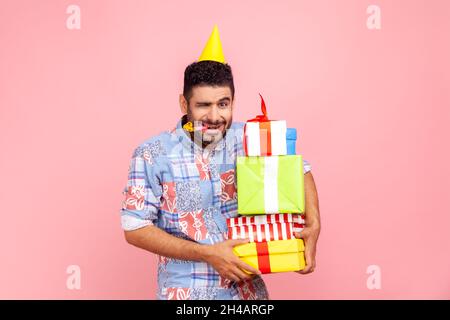 Junger erwachsener lustiger bärtiger Mann mit runzelndem Gesicht, Blick auf die Kamera und Augenzwinkern, bläst Partyhorn, hält Stapel von Geschenken, Geburtstag. Innenaufnahme des Studios isoliert auf rosa Hintergrund. Stockfoto