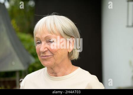 Nachdenkliche ältere Dame, die ihre Lippen mit einem nachdenklichen Lächeln stäupelt, während sie leise mit einem wehmütigen Ausdruck zur Seite starrt Stockfoto