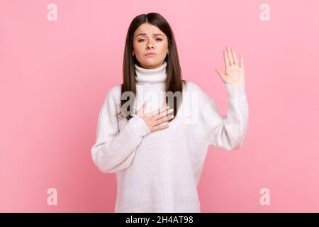 Porträt einer ehrlichen jungen Frau, die mit der Hand auf der Brust und den Fingern nach oben steht und Loyalität verspricht, in einem weißen Pullover im lässigen Stil. Innenaufnahme des Studios isoliert auf rosa Hintergrund. Stockfoto