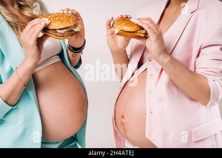Zwei Schwangere in Anzügen mit Hamburgern in den Händen auf grauem Hintergrund. Stockfoto
