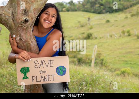 Junge Erwachsene lateinische Mädchen hält Zeichen für die Rettung des Planeten in einem tropischen Ort. Platz für Text. Brünette Frau umarmt Pflanze. Stockfoto