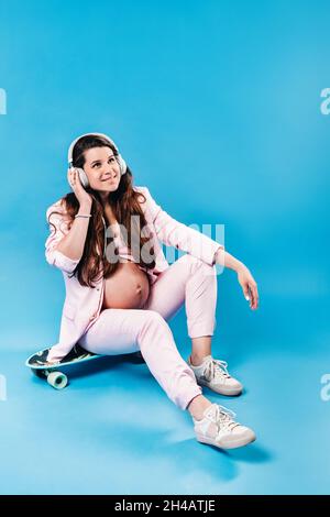 Eine Schwangere im pinken Anzug sitzt mit Kopfhörern auf einem Skateboard und hört Musik auf blauem Hintergrund. Stockfoto