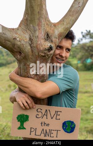 Der Mensch fragt, den Planeten zu retten. Ein junger erwachsener latein hält ein Schild, auf dem er darum bittet, den Planeten zu retten, während er einen Baum umarmt. RETTE DEN PLANETEN. Stockfoto