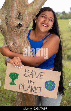 Vertikales Foto einer Frau, die um die Rettung des Planeten bittet. Junge lateinische Erwachsene Frau hält ein Schild mit der Bitte, den Planeten an einem tropischen Ort zu retten. Rette Die Erde Stockfoto